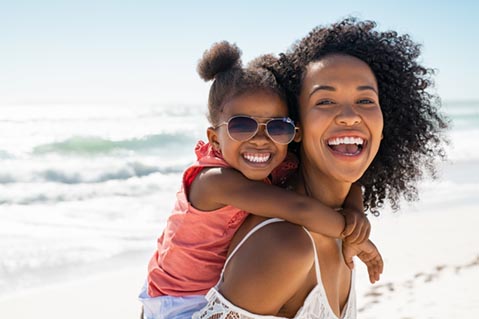 A child wearing sunglasses at the beach while being carried by her mother